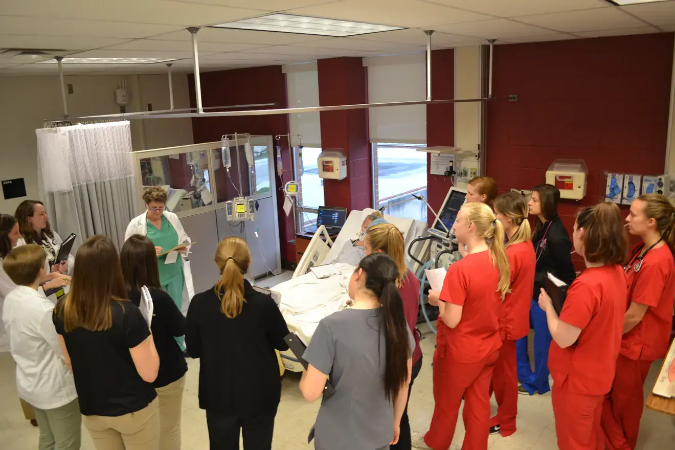 Learners standing around a simulation lab with a patient on a bed in the middle of the huddle.