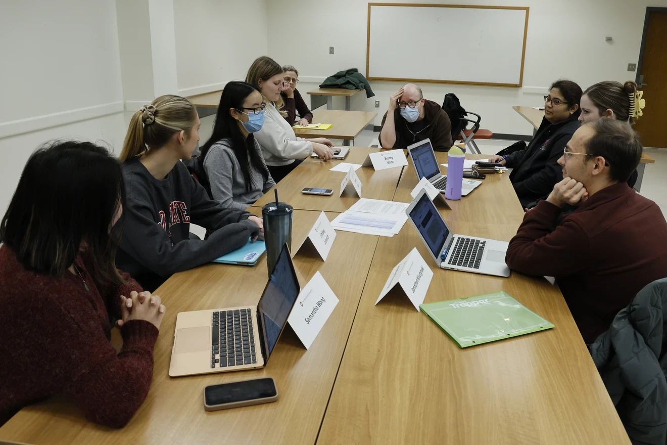 A group of individuals seated around a rectangular table in a classroom setting. Each person has a laptop open in front of them, and there are papers and notebooks on the table as well. The focus is on collaboration or a group discussion, indicated by the arrangement of the seating and the attentive postures. A whiteboard is visible in the background, but it is blank.