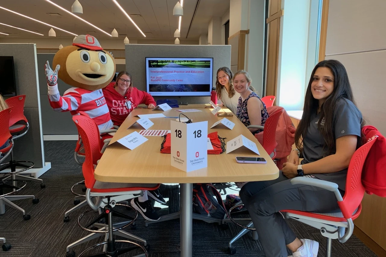 The image shows a group of individuals seated at a table in a well-lit room with large windows. One person is dressed in an Ohio State Buckeyes mascot costume, complete with a large head depicting a character with an “O” on its cap and sports attire. The mascot is posing with one hand raised in a greeting gesture. In front of the individuals are laptops, papers, and what appears to be study materials. There’s also a monitor on the table displaying materials.