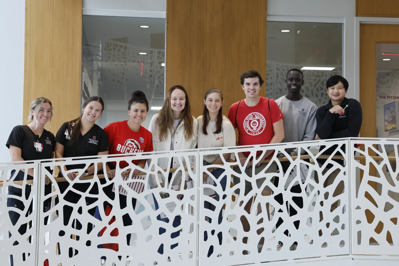 A group of seven individuals stands behind a decorative railing with cut-out patterns, possibly on a balcony or an indoor overlook. The faces of the individuals are pixelated for privacy. The group appears diverse in terms of clothing style, with some wearing casual attire and others in more formal or branded apparel. The background suggests a public or communal space