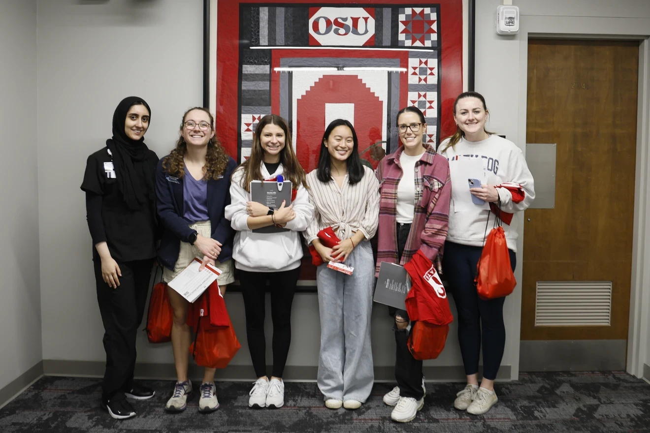 The image shows a group of seven individuals standing in front of a large Ohio State University (OSU) banner. The banner features the OSU logo, an image of a building, and various patterns in red, black, and white. Each person is holding informational materials and red bags