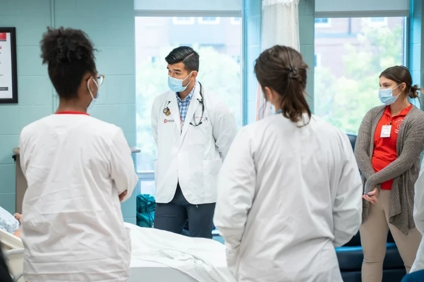 A group of medical professionals or students, dressed in white coats and face masks, gather around a hospital bed in a clinical setting. One individual, likely a doctor or instructor, leads a teaching session, emphasizing hands-on medical education. The background includes medical equipment and privacy curtains, reinforcing the hospital environment.