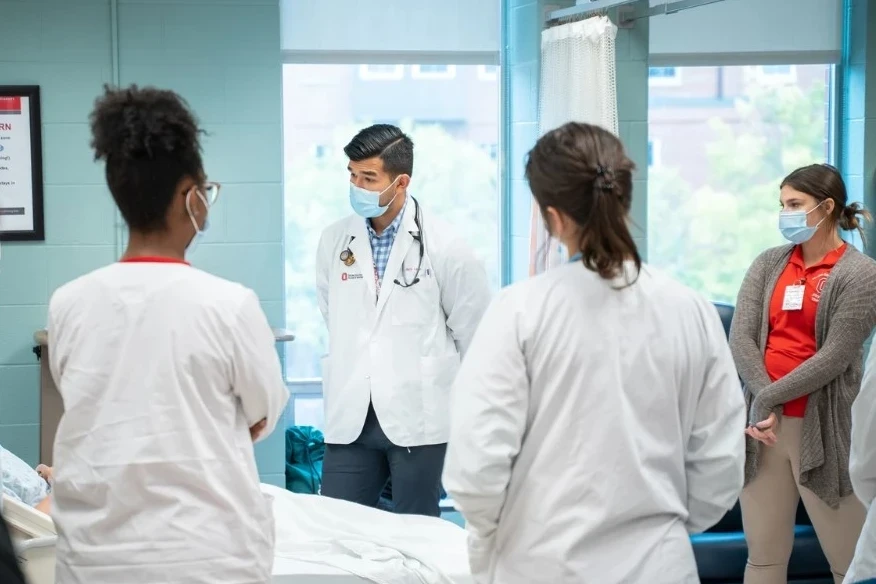 A group of healthcare professionals and students are gathered in a clinical or classroom setting. They are wearing medical attire, including white coats and scrubs, and holding clipboards or binders. One individual in green scrubs has a stethoscope around the neck and appears to be speaking or explaining something to the group. The background shows a wall clock and a doorway, suggesting an indoor educational or hospital environment.