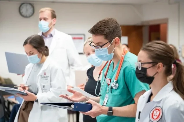 A group of healthcare professionals and students are gathered in a clinical or classroom setting. They are wearing medical attire, including white coats and scrubs, and holding clipboards or binders. One individual in green scrubs has a stethoscope around the neck and appears to be speaking or explaining something to the group. The background shows a wall clock and a doorway, suggesting an indoor educational or hospital environment.
